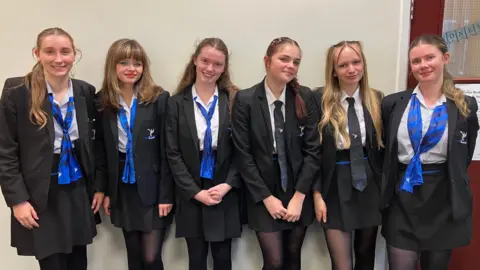 BBC Six teenage girls in black school uniforms which have a blue and white crest in all smiling and looking at the camera.