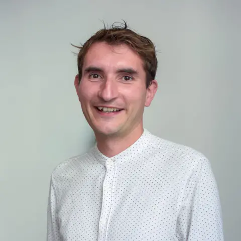 The Data City A man smiles at the camera. He is wearing a collarless white shirt buttoned up. The shirt has small dots on it. The man has brown hair and is clean-shaven.