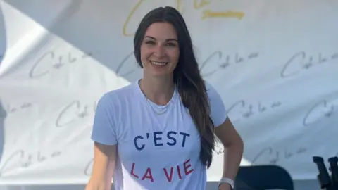 Deanna Newman A woman with long brown hair is wearing a t-shirt that says 'C'est La Vie'.