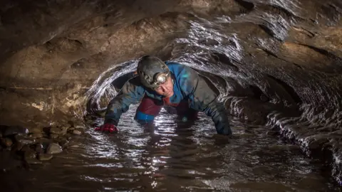 Gary Douthwaite The photo shows a person wearing a helmet with a headlamp crawling through a narrow, wet cave passage, surrounded by rocky walls and water on the ground.