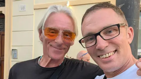 Phil James Singer Paul Weller is dressed in a black t-shirt and orange sunglasses. He has long white hair and has his arm around a man in a white-shirt and black glasses. They are standing outside a cream building with a wooden door.