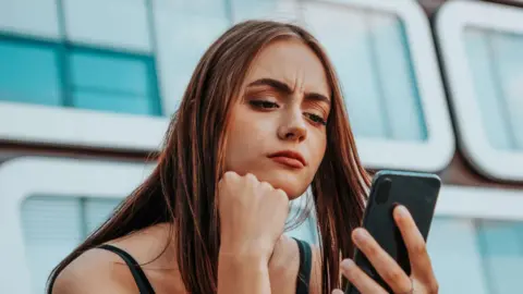 Getty Images A young woman, looking concerned, looks at here smartphone.