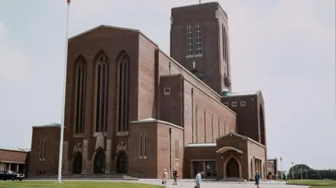 Historic England Archive An austere brown brick cathedral with tall arched windows and sections of several different heights, including a tower. There is an area of green grass with a flag pole in front of the cathedral and a black car parked to the left. Six older people can be seen walking in the foreground. The sky looks murky and overcast, a muted blue/grey with big clouds.