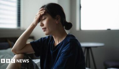 A man with grey hair on the sides of his head and glasses smiles at the camera. He is wearing a grey suit jacket.