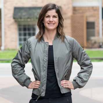 Woman standing outside with her hands on her hips looks at the camera. She is wearing a black T-shirt and a green jacket. 