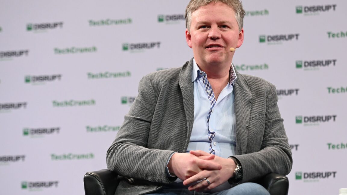 A man in a suit jacket sits at a TechCrunch speaking event