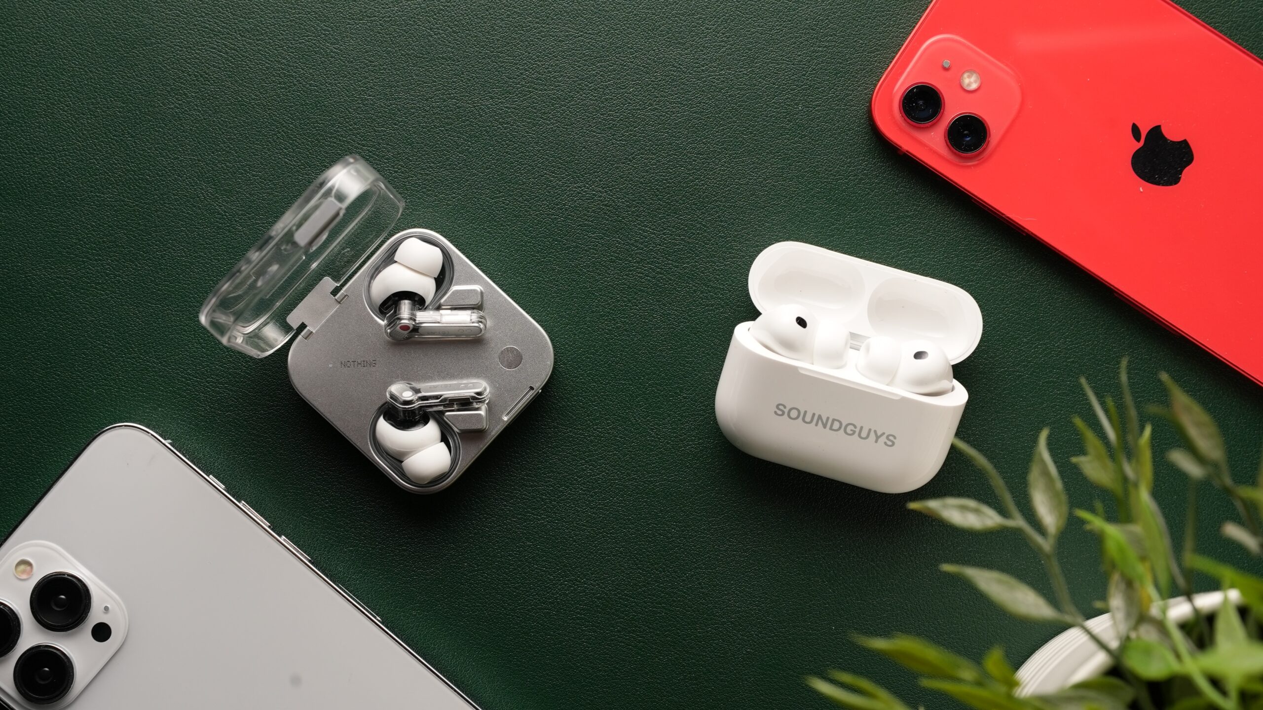 The AirPods Pro 3 and the Nothing Ear (3) sit side-by-side against a dark green backdrop, surrounded by iPhones and a plant.