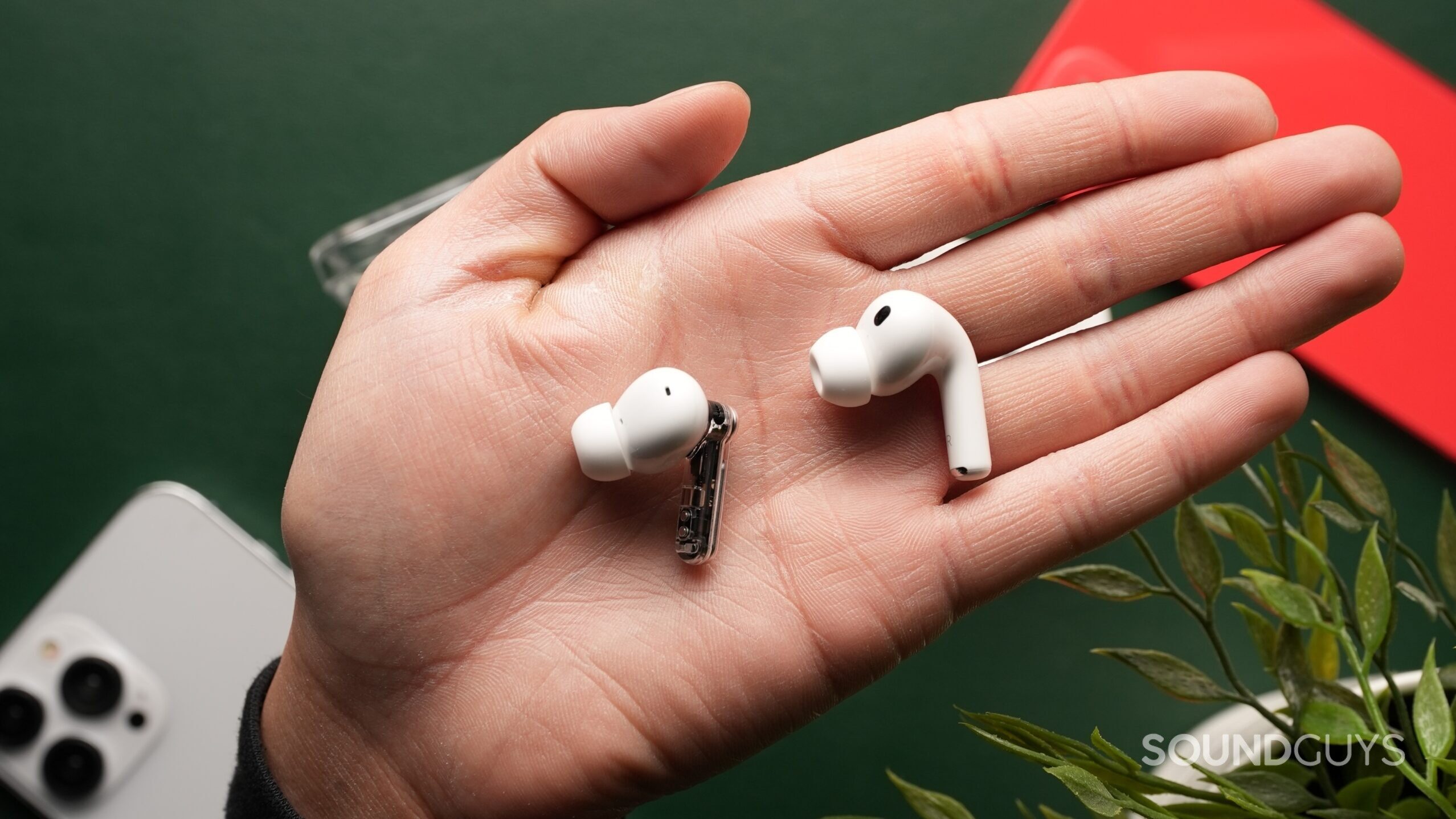 The AirPods Pro 3 and the Nothing Ear (3) sit out of their cases side-by-side on a palm against a dark green backdrop, surrounded by iPhones and a plant.