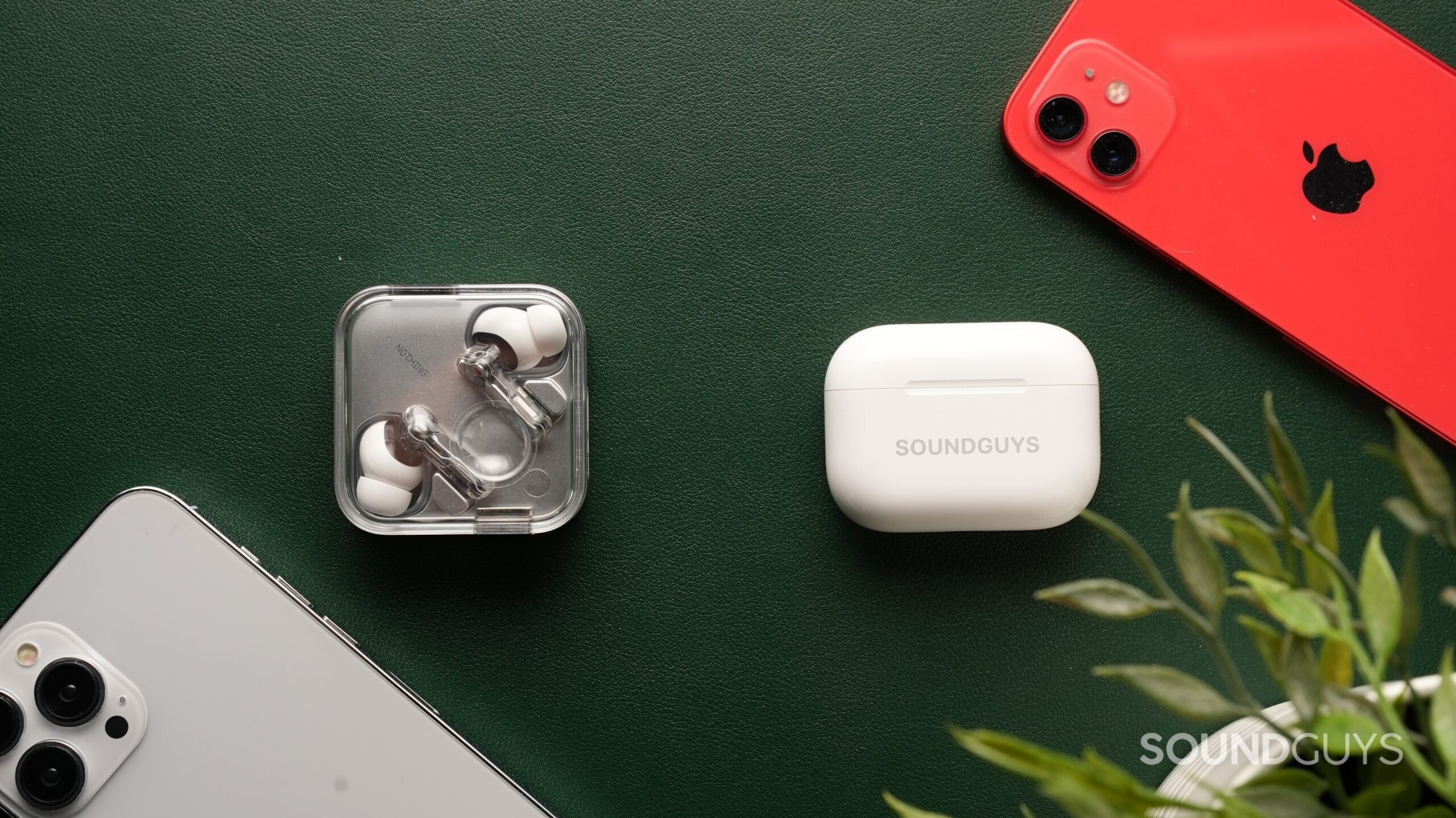 The AirPods Pro 3 and the Nothing Ear (3) sit side-by-side against a dark green backdrop, surrounded by iPhones and a plant.