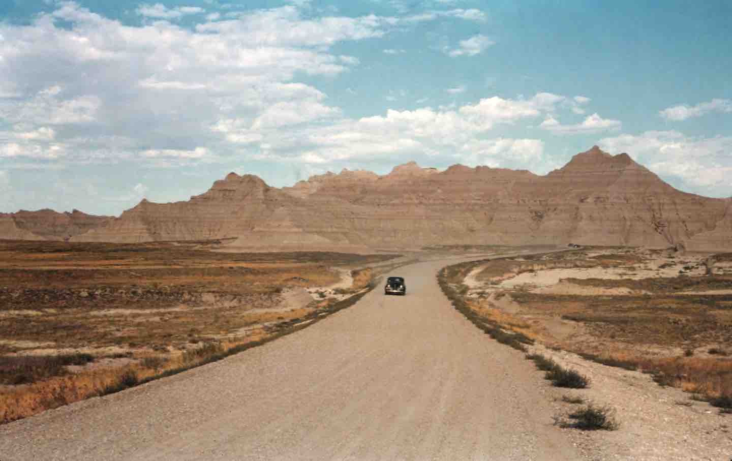 A 35mm film photo shows an automobile making its way down an empty dirt road in Badlands National Park, 1940.
