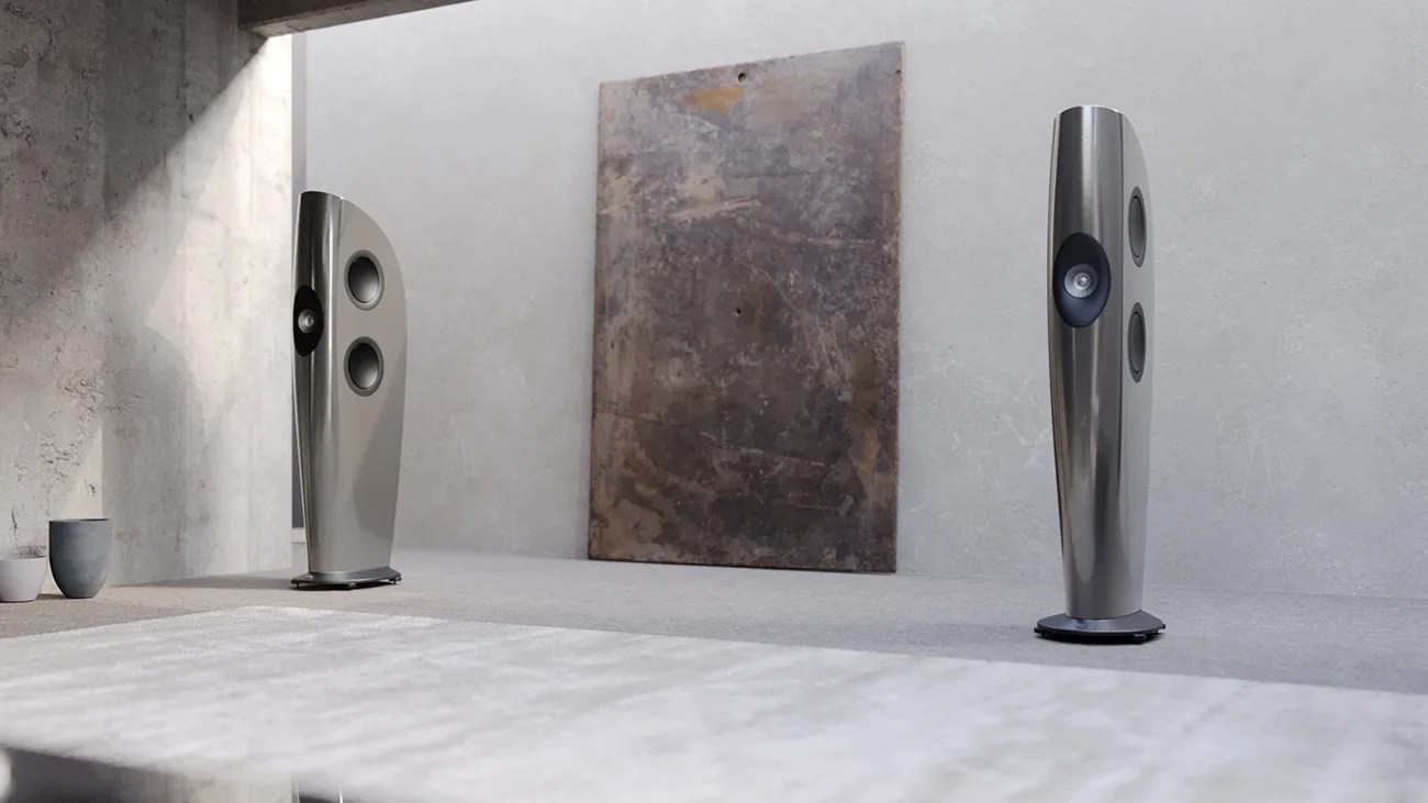 Pair of tall, sleek, metallic floor-standing speakers in a minimalist room with concrete walls and a large abstract wall art.