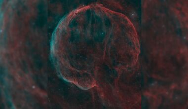 A deep space nebula scene showing a bubble-like shell of cosmic dust and gas shining red and blue against a black sky peppered with bright stars.