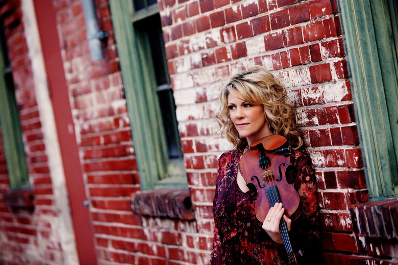 A middle aged white woman with blonde curly hair and wearing a red and black dress stands against a red brick wall, a fiddle tucked under her chin.