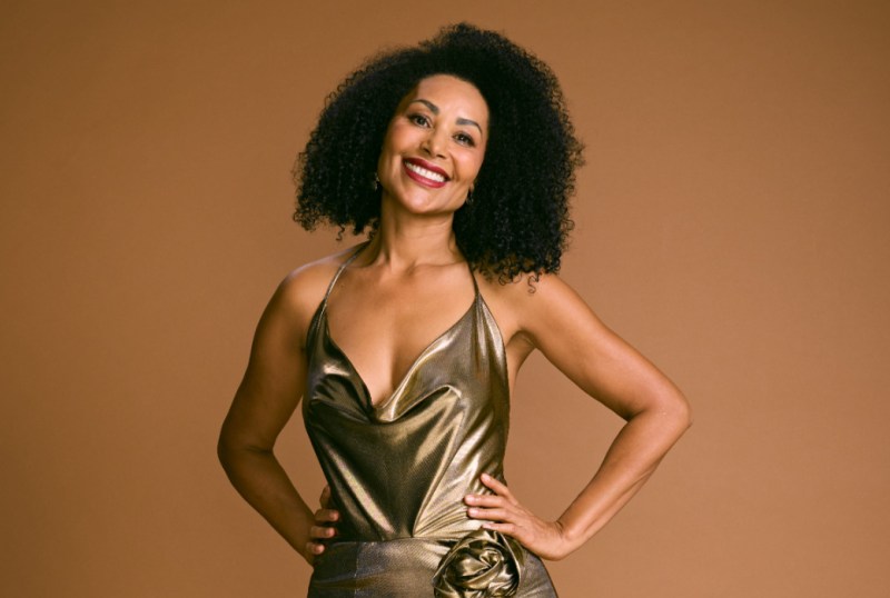Smiling woman with curly hair wearing a shimmering gold dress, posing confidently against a warm brown background.