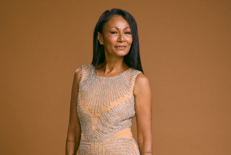 A confident woman with long black hair poses in a sparkling, intricately beaded dress against a warm brown background, showcasing elegance and poise.
