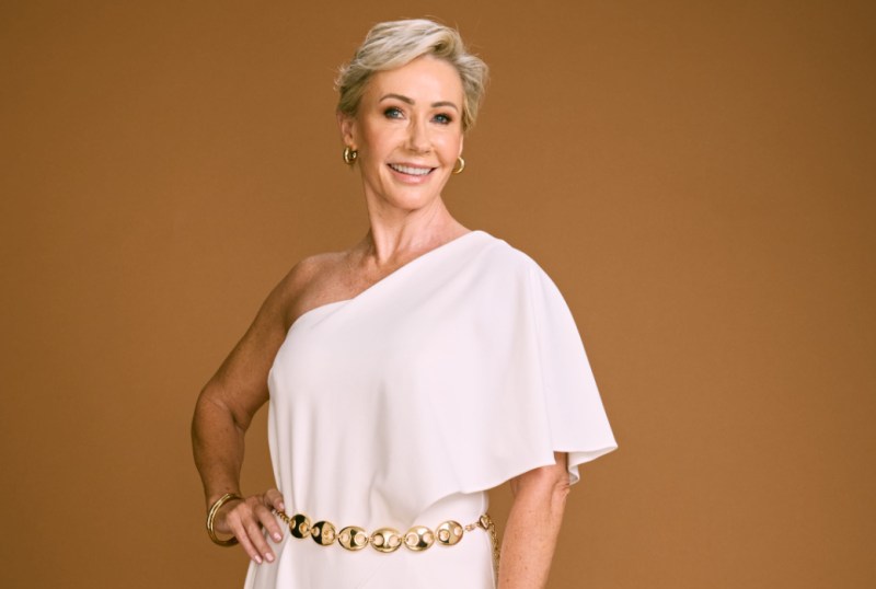 Smiling woman in a stylish white one-shoulder dress with a gold belt, posing confidently against a warm brown background. Her short blonde hair and gold accessories complement her elegant look, showcasing a modern fashion statement.