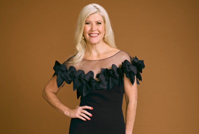 Smiling woman with long blonde hair wearing a black dress with a floral ruffle design on the neckline, standing against a warm brown background.