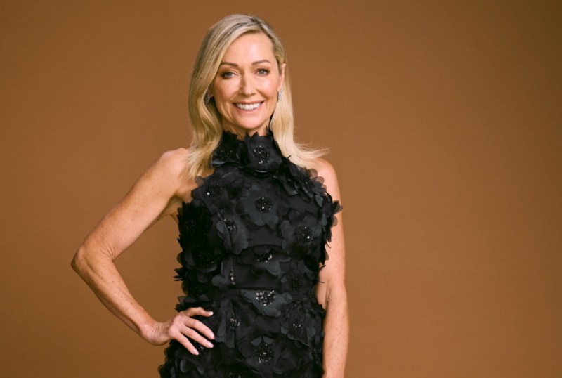 Smiling woman in a black floral dress poses against a brown background, showcasing a stylish and elegant look.