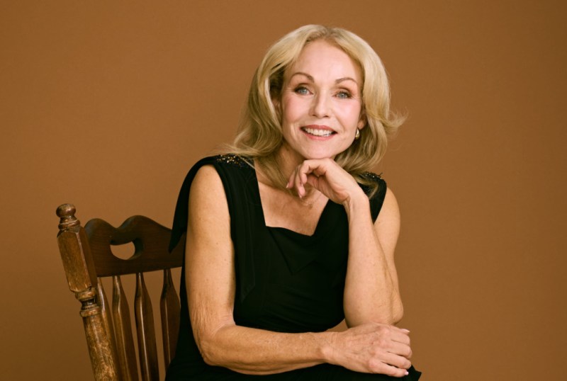Smiling woman with long blonde hair wearing a black dress, seated on a wooden chair against a warm brown background. The image captures a confident and approachable expression, perfect for showcasing elegance and charm.