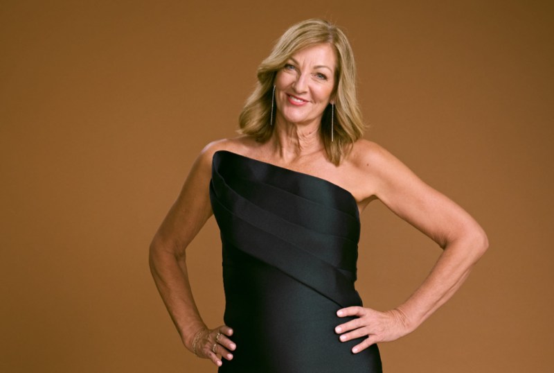 Smiling woman in a strapless black dress, posing confidently with hands on hips against a warm brown background, showcasing elegance and style.