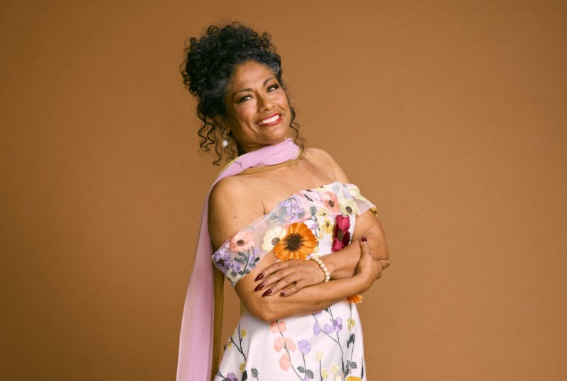 Smiling woman with curly hair wearing a floral off-shoulder dress and a pink scarf, posing confidently against a warm brown background.