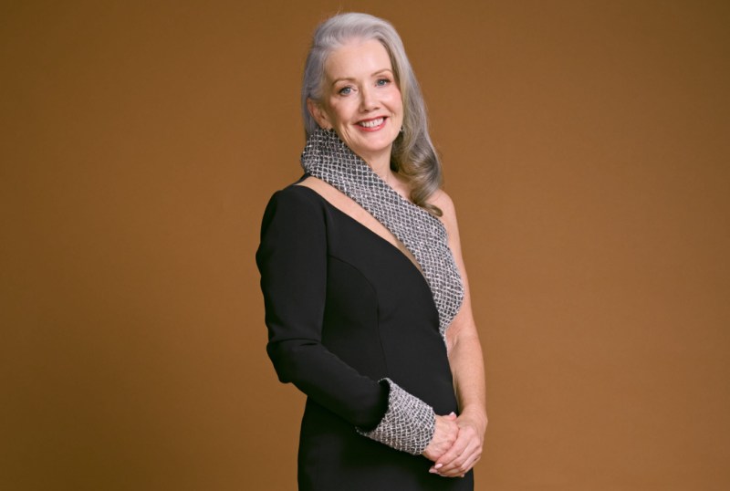 Elegant woman in a sophisticated black dress with a unique silver-accented neckline and sleeve, posing confidently against a warm brown background.