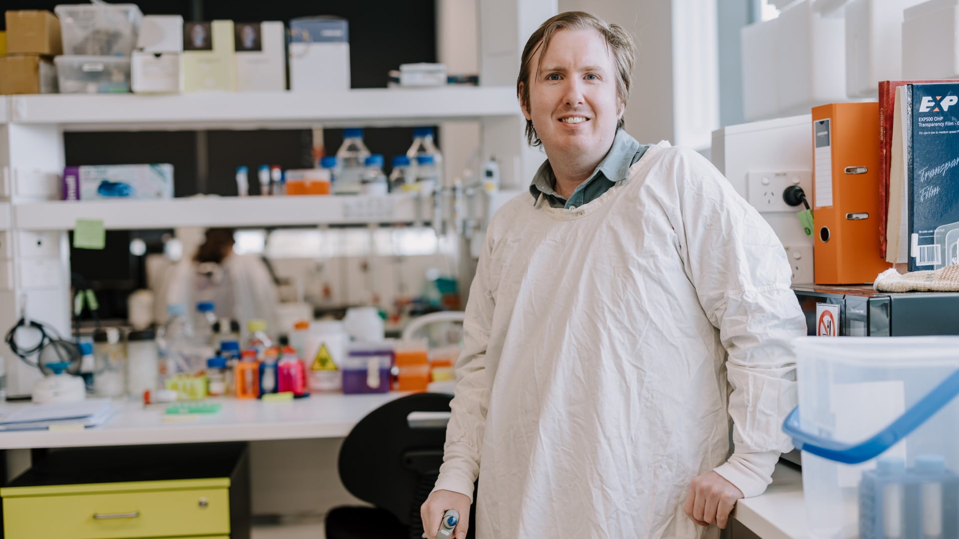 Luke McAlary, in a white lab coat, leans against a bench in a laboratory. Photo: Michael gray