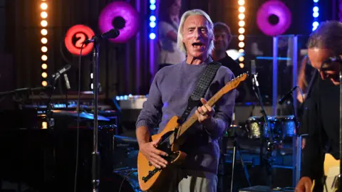 Singer Paul Weller has long white hair, a dark blue jumper, grey trousers and is playing a wooden electric guitar. There is a microphone on a stand close by him. A drummer can be seen in the background with his kit and another man dressed in black playing a guitar. There is other music equipment in the performance area and bright neon lights on the wall. One is in the shape of the number 2.