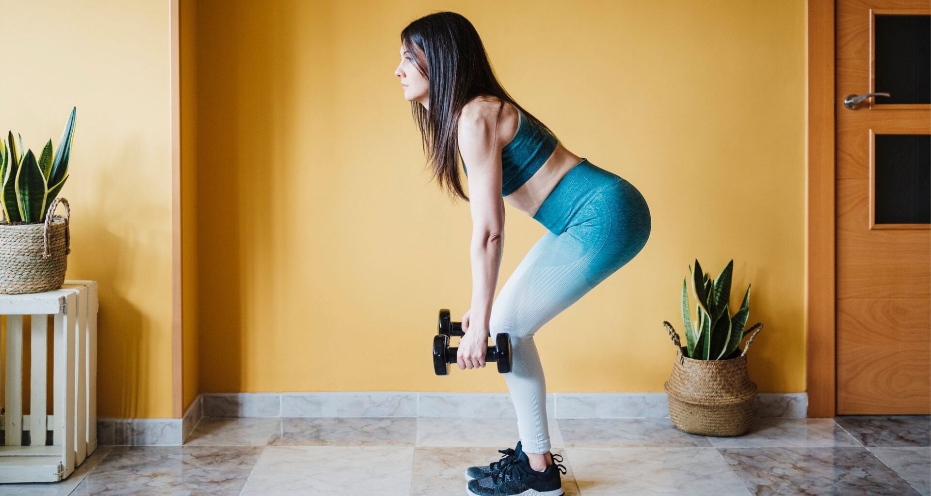 woman in leggings and crop top holding two dumbbells half way through a deadlift. she's sideways to the camera with a yellow wall and a couple of plants behind her