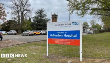 The main sign at the entrance of Hellesdon Hospital on the outskirts of Norwich. It states: "Norfolk and Suffolk NHS Foundation Trust" on the white section; "Welcome to Hellesdon Hospital" in the blue section, and some indecipherable text in the red section. Around it is lawn, trees, a car park with cars parked and a tower of the hospital structure.
