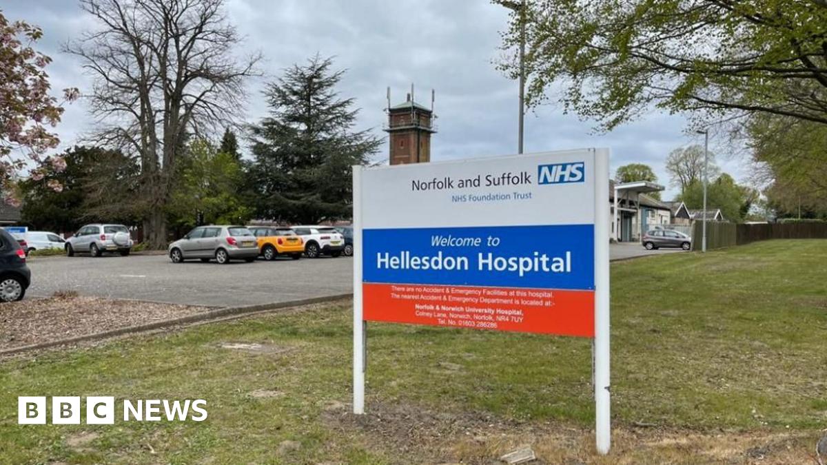 The main sign at the entrance of Hellesdon Hospital on the outskirts of Norwich. It states: "Norfolk and Suffolk NHS Foundation Trust" on the white section; "Welcome to Hellesdon Hospital" in the blue section, and some indecipherable text in the red section. Around it is lawn, trees, a car park with cars parked and a tower of the hospital structure.