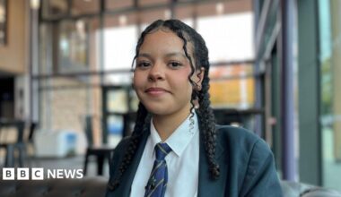 Sam Coton wears a navy polo shirt with the 'Better Me, Better You' logo on it. He is sitting in the school theatre bar with floor to ceiling glass windows, you can see tables and chairs behind him. He is smiling at camera.