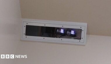 The Oxevision technology - which is an infrared camera - placed near the ceiling in the corner of a bedroom. It is in a white rectangle box and small white lights can be seen at one end.