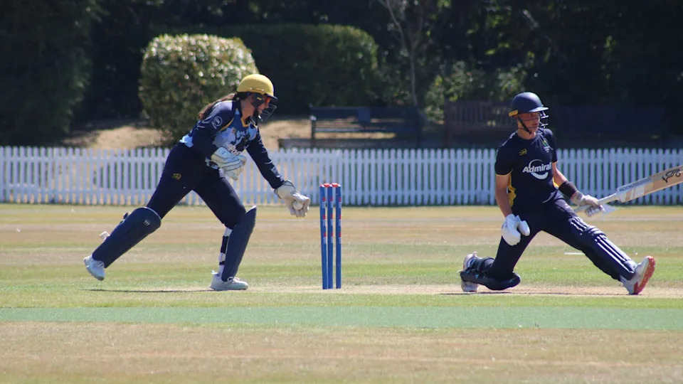 Libby, with a long brown ponytail under her yellow helmet takes the ball in her left glove as she prepares to whip off the bails