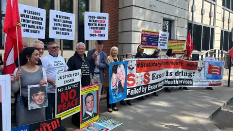 Richard Daniel/BBC Several campaigners stand outside a building holding placards and signs calling for changes in how mental health services are handled. 