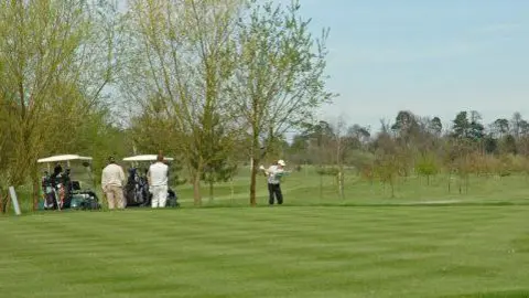 Geograph A golfer wearing a white hat, pale jumper and dark trousers, playing a drive at a golf course. A number of other people are standing behind the officer and watching.