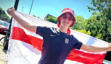 A young man wearing sunglasses, a blue football shirt and a bucket hat with his arms outstretched, hold an England flag behind him
