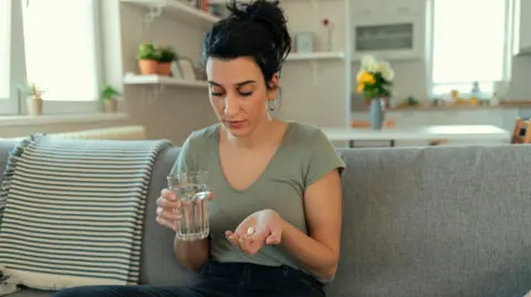 Getty Images Young woman is sat with a glass of water in one hand and a white tablet in the open palm of the other. She is looking down so you can't see her eyes and her hair is tied up. She is sat on a grey sofa in what looks like an open plan living room with the kitchen behind her. 