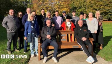 People stood in grass area around wooden bench. Two men sat on a bench. There are trees in the background.
