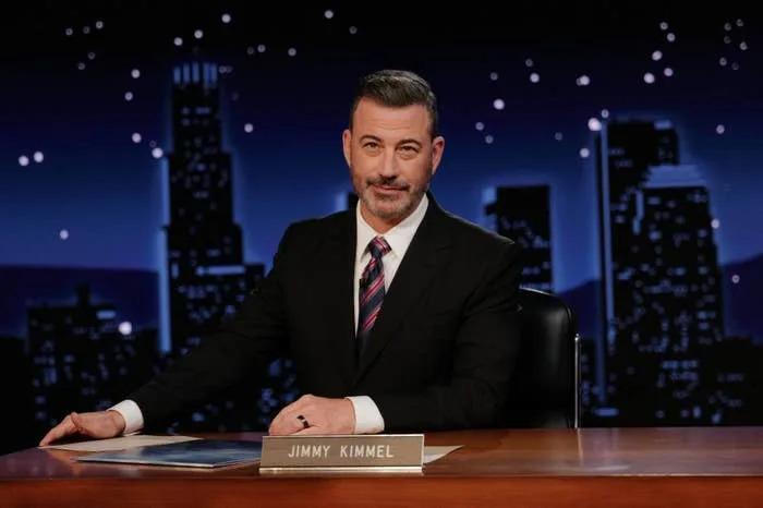A man sits confidently at a talk show desk with a cityscape backdrop, wearing a suit and tie, in a nighttime TV show setting