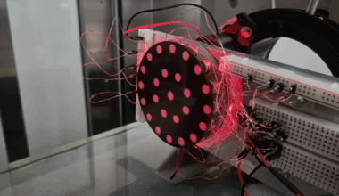 A vertically-mounted black disk with a concentric pattern of reflective disks is illuminated under a red light. A large number of copper wires run away from the the disk to a breadboard.