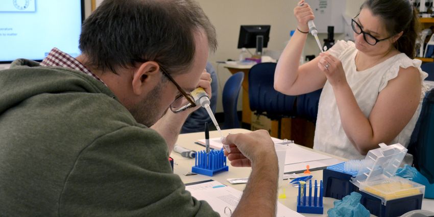 Photograph of two teachers, a woman in a white shirt with dark hair and glasses and a man with short dark hair in a green sweatshirt, at a table holding small plastic tubes and pipettes to perform DNA extraction.