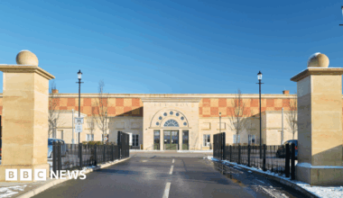Front of a modern building. It has an arched reception area and there are two stone columns in the foreground indicating the entrace. It is yellow and orange coloured. At the centre of the picture is a road with markings.
