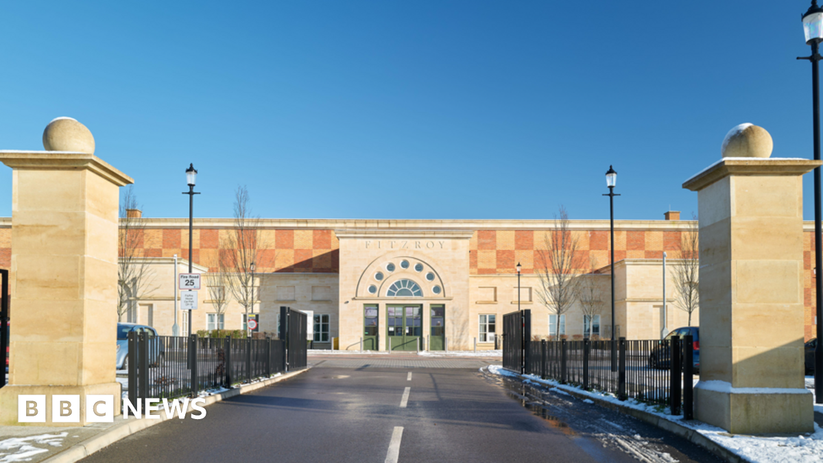 Front of a modern building. It has an arched reception area and there are two stone columns in the foreground indicating the entrace. It is yellow and orange coloured. At the centre of the picture is a road with markings.
