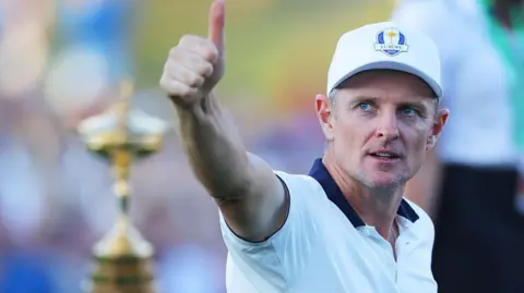 Getty Images Ryder Cup European golfer Justin Rose wearing a white short sleeve top and a Ryder Cup hat. His right arm is pointing towards the camera with his thumb up. The Ryder Cup trophy is in silhouette in the background.