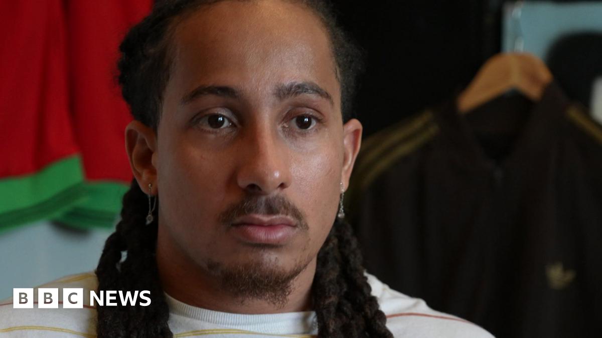 A head-and-shoulders image of Tyler Attwood who is looking serious. He has long dark brown hair styled in dreadlocks. There are sports tops on hangers behind him.
