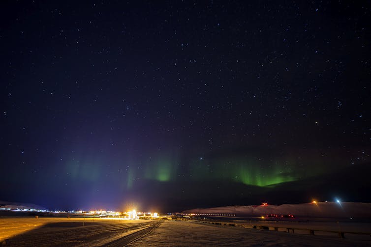 Several buildings on an icy plain, with green lights in the sky above.