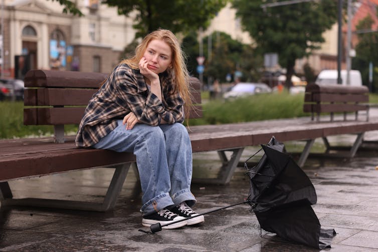 A young woman sits alone on a bench outside, gazing thoughtfully.