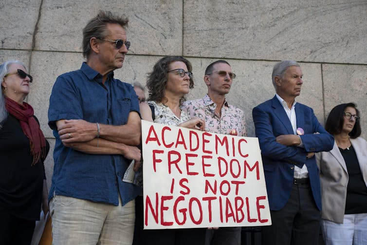 Several adults stand together and look serious, holding a sign that says 'Academic freedom is not negotiable.'