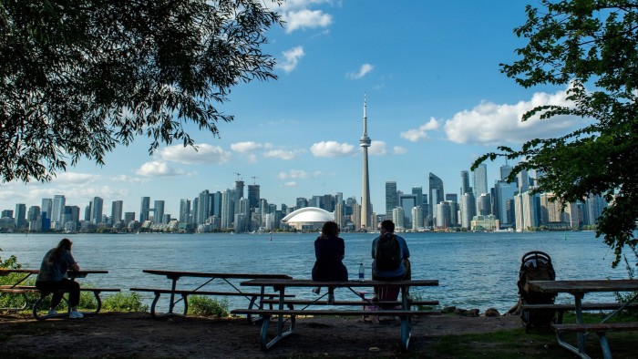 People sit on benches under trees, looking across Lake Ontario at the CN Tower and Toronto skyline on a sunny day.